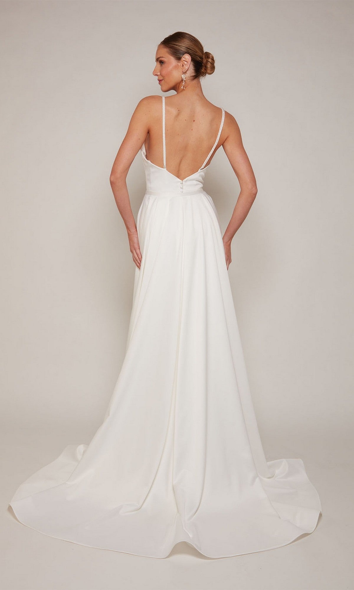 Woman in the Alyce 7090 long formal dress—a white satin gown with a low back, long train, and detachable overskirt—stands against a plain background, looking over her shoulder.